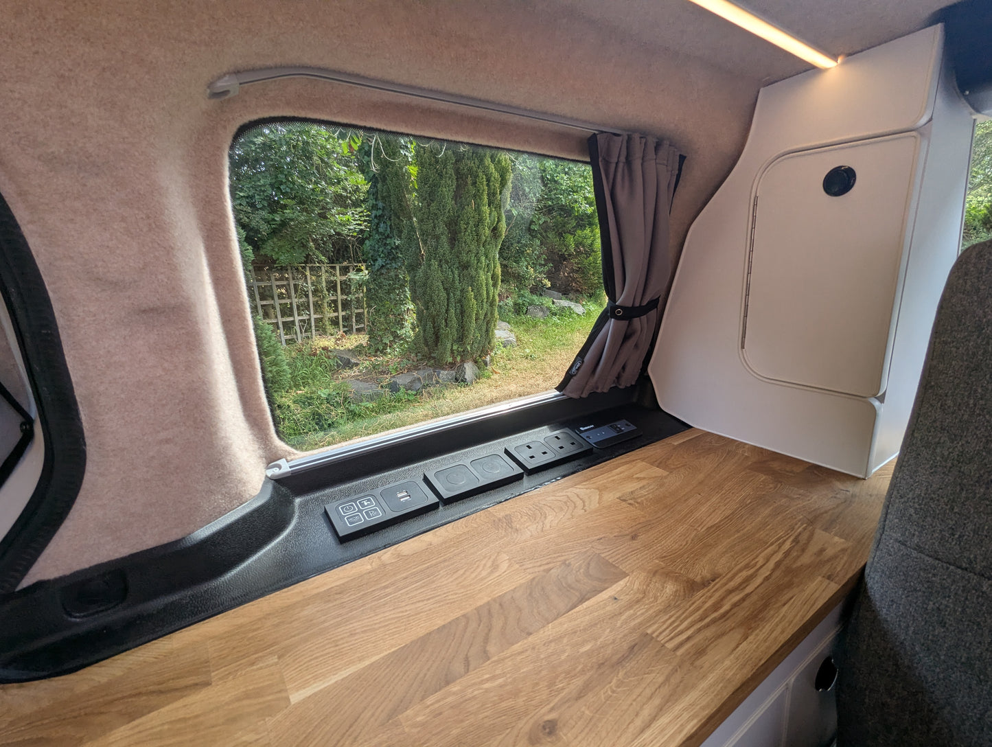 Interior view of a vw id buzz camper van showing a wooden workbench, beige wall panels, a window with a curtain, and an outdoor garden view.