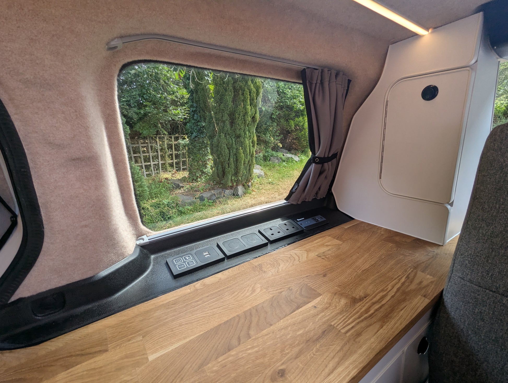 Interior view of a vw id buzz camper van showing a wooden workbench, beige wall panels, a window with a curtain, and an outdoor garden view.