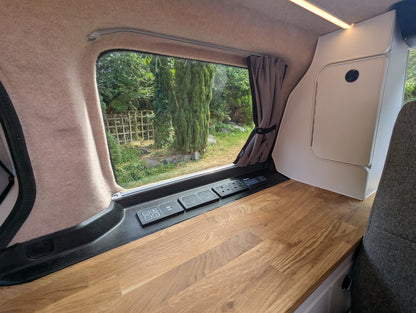 Interior view of a vw id buzz camper van showing a wooden workbench, beige wall panels, a window with a curtain, and an outdoor garden view.