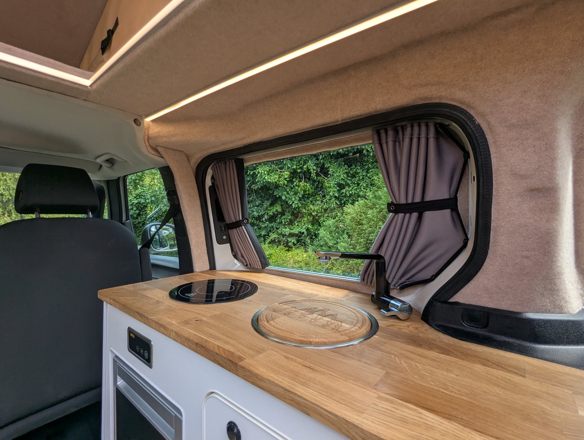 A compact kitchenette installed inside a vw id buzz camper van, featuring a light wood countertop with a built-in black cooktop and glass lid, white storage cabinets, gray curtains, and a window looking out to green foliage.