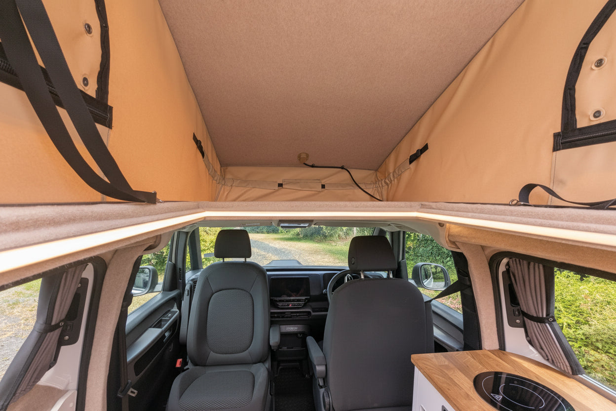 The interior of a vw id buzz camper van with a raised beige canvas roof, showing the front seats, windows, and a small wood-topped kitchen area in the lower right.