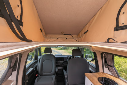 The interior of a vw id buzz camper van with a raised beige canvas roof, showing the front seats, windows, and a small wood-topped kitchen area in the lower right.