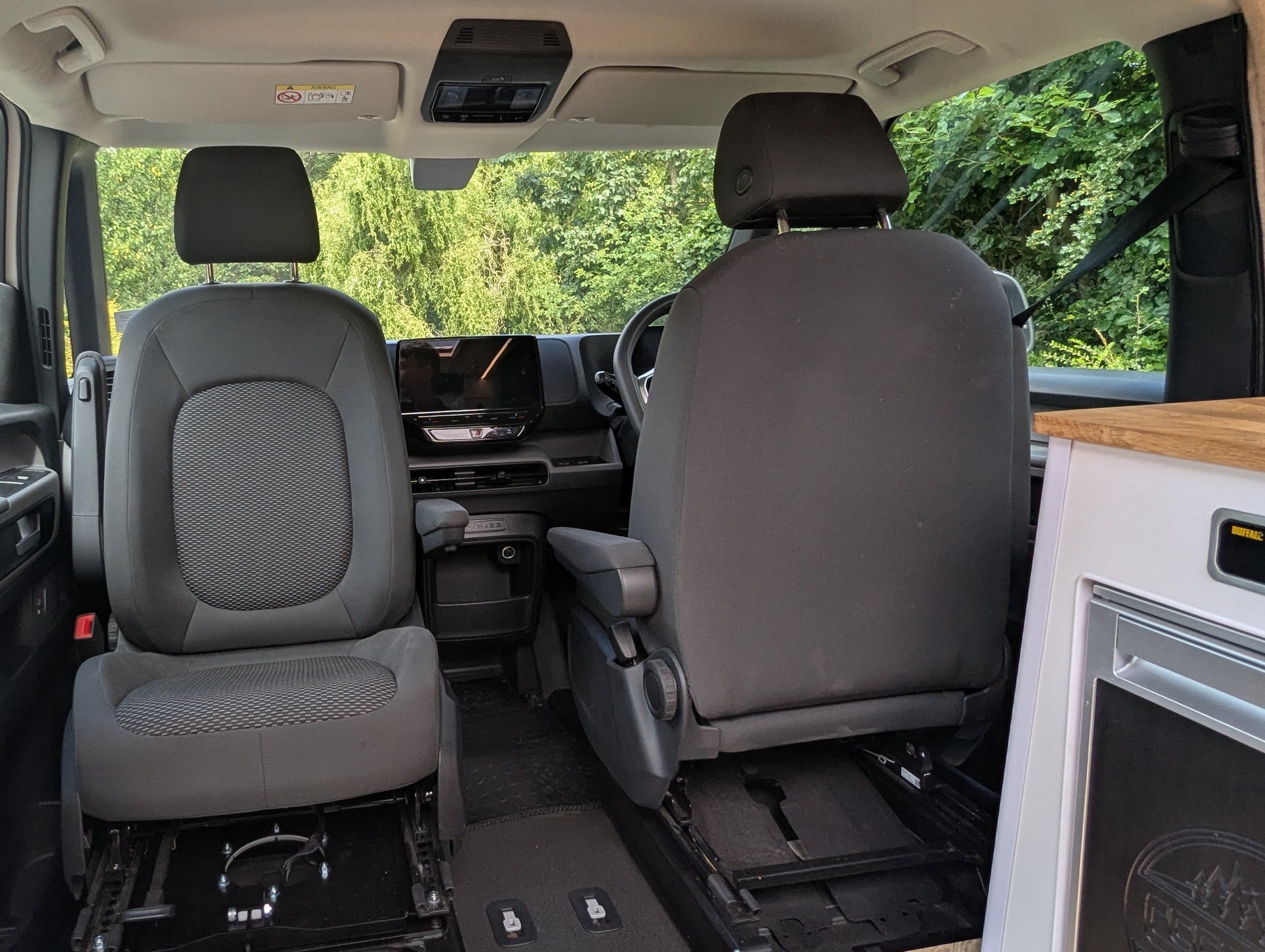 The interior of a Volkswagen ID. Buzz electric campervan showing the spacious swivel seat with a wood-look floor, and a compact kitchenette on the right side.