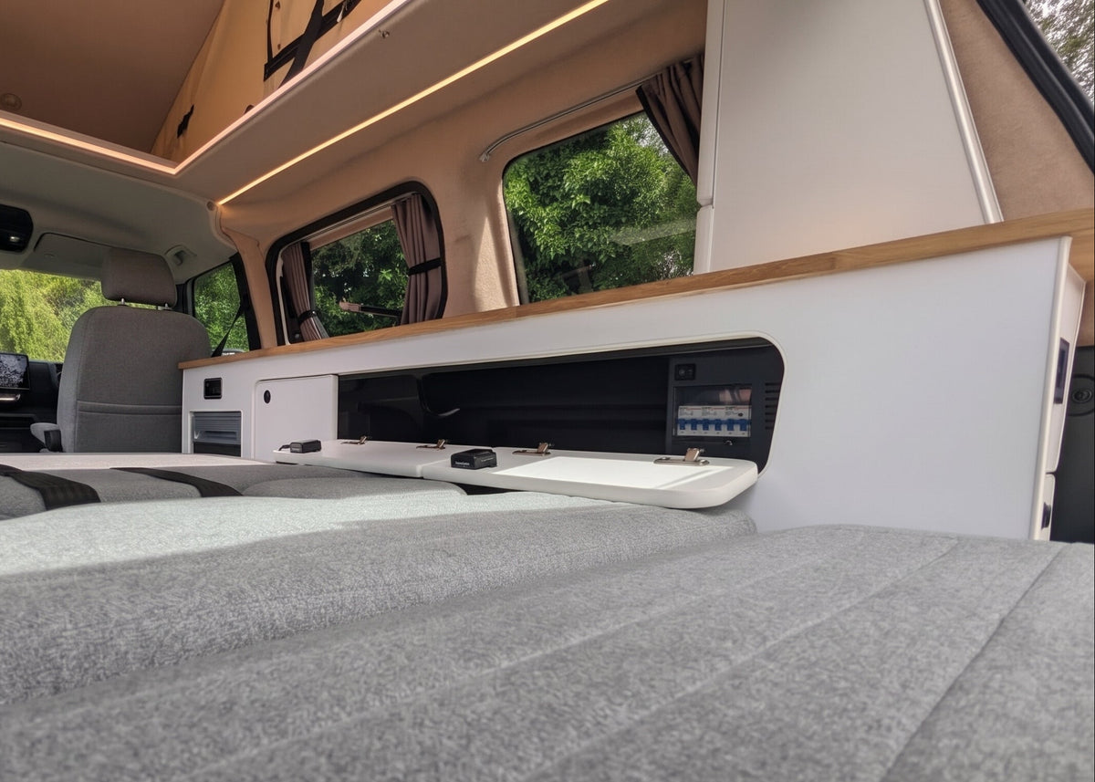 A low-angle view inside a camper van showing a gray upholstered bed or seating surface in the foreground, with wood trim, windows, and interior cabinetry behind it.