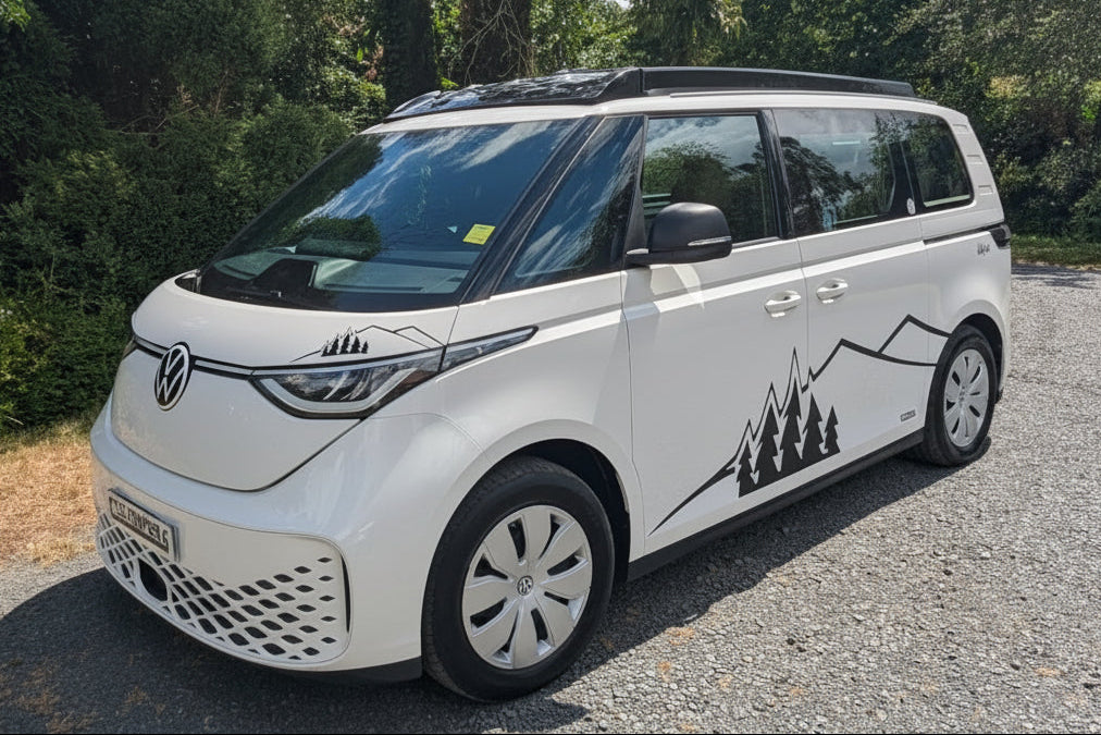 A white Volkswagen ID. Buzz electric campervan parked on a gravel path outdoors, surrounded by green trees and bushes under a partly cloudy sky.