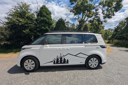A white electric Volkswagen ID. Buzz campervan with a black mountain and pine tree graphic decal on the side, parked on a gravel path outdoors with trees and a blue sky in the background.