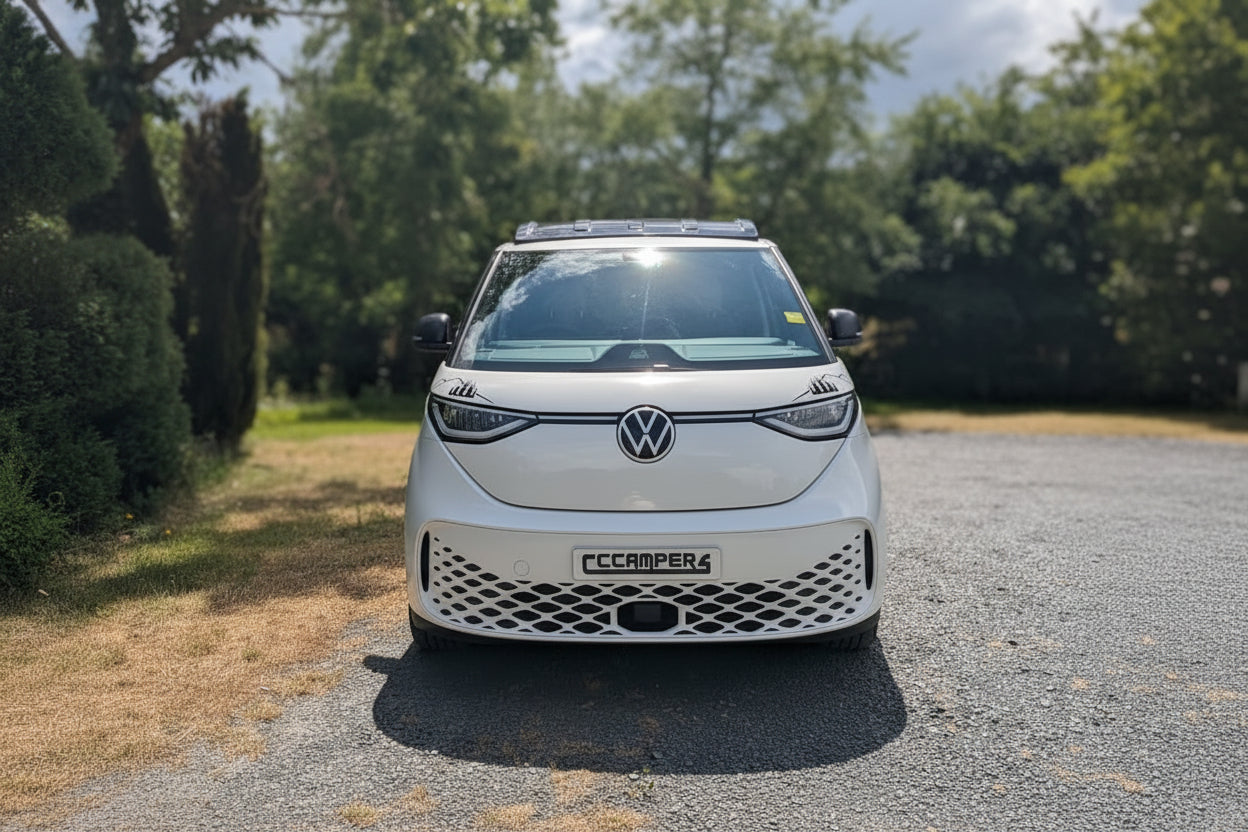 A white Volkswagen ID. Buzz electric campervan parked on a gravel path outdoors with trees and a cloudy sky in the background.