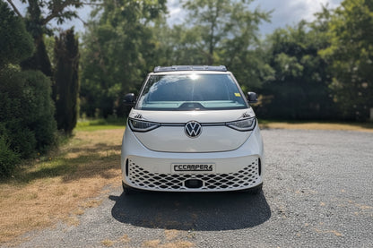 A white Volkswagen ID. Buzz electric campervan parked on a gravel path outdoors with trees and a cloudy sky in the background.