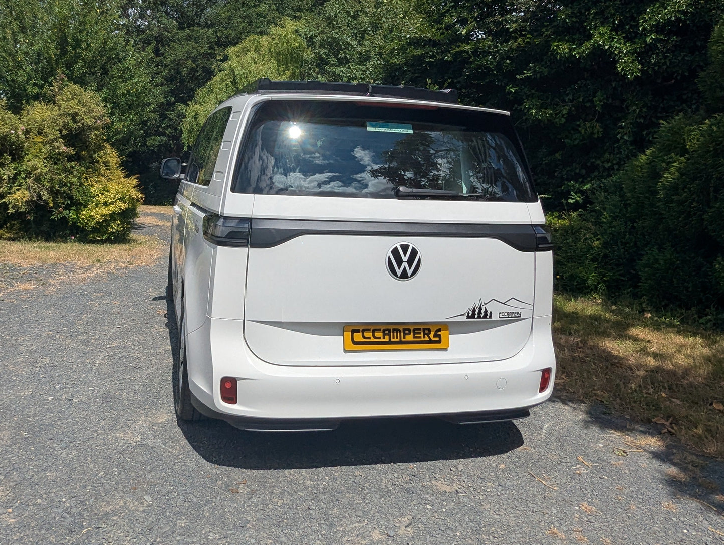 A white Volkswagen ID. Buzz electric campervan parked on a gravel path outdoors, photographed from the rear. The vehicle features a yellow 'CCAMPER4' license plate and a black horizontal trim line across the tailgate, with green trees and bushes in the background.