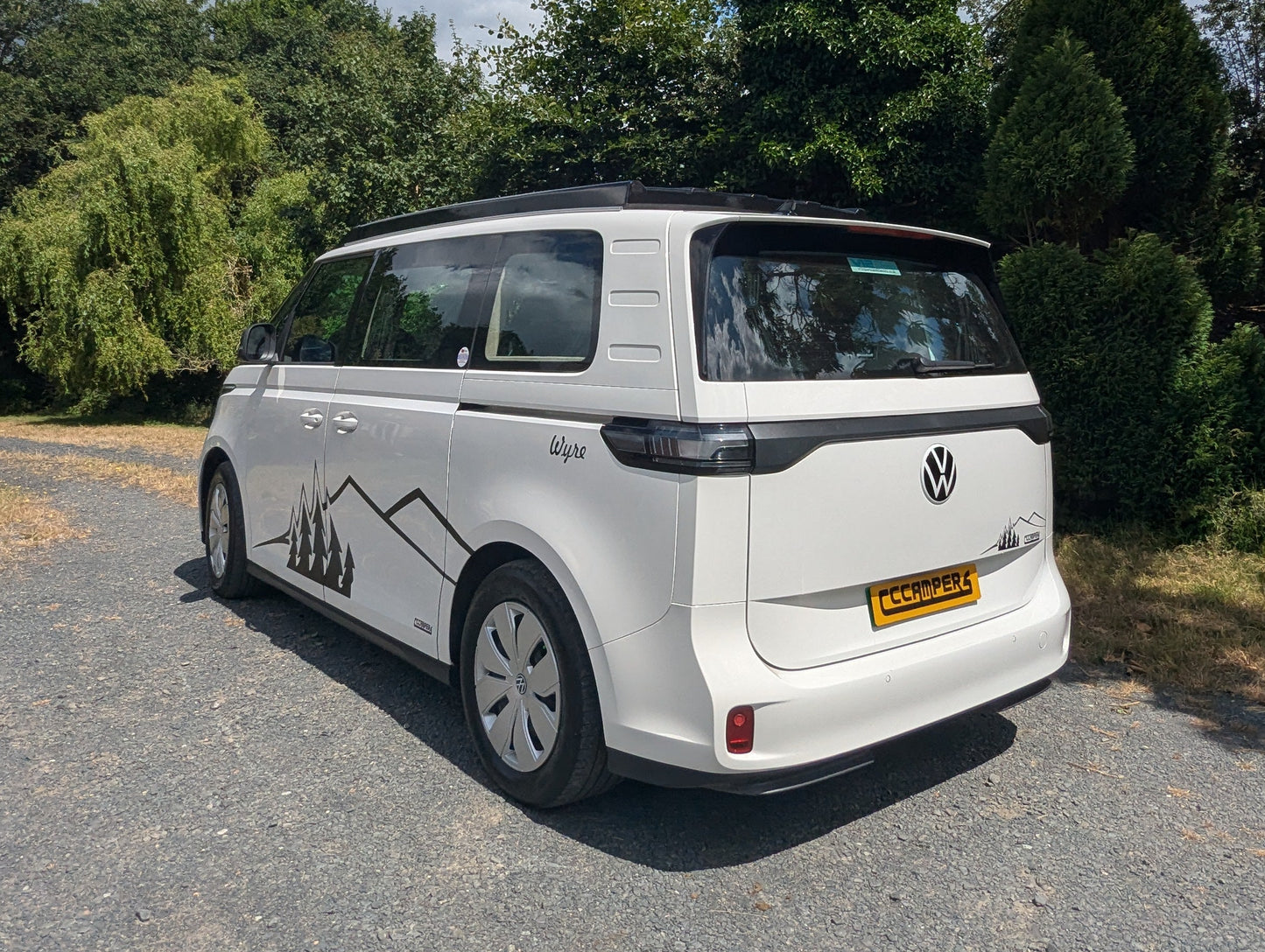 A white Volkswagen ID. Buzz campervan parked on a gravel path outdoors, surrounded by trees and bushes under a blue sky with clouds.