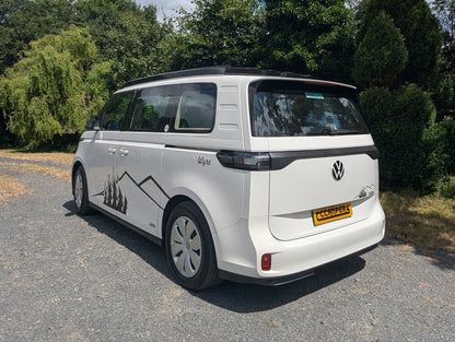 A white Volkswagen ID. Buzz campervan parked on a gravel path outdoors, surrounded by trees and bushes under a blue sky with clouds.