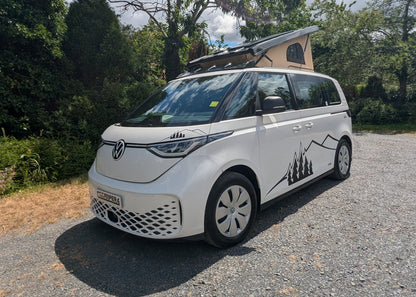 A white Volkswagen ID BUZZ electric campervan with a rooftop pop-up tent, parked on a gravel driveway surrounded by trees and a blue sky.
