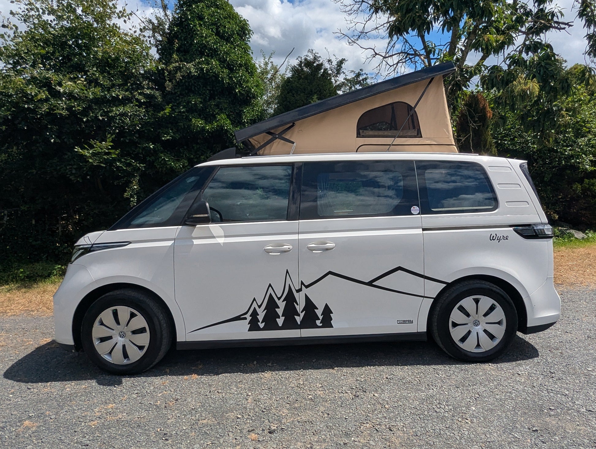The Wyre by CCCAMPERS — white Volkswagen ID. Buzz electric campervan with Stargaze rear-elevating pop-top roof extended, parked outdoors with signature mountain and pine tree graphic decal.