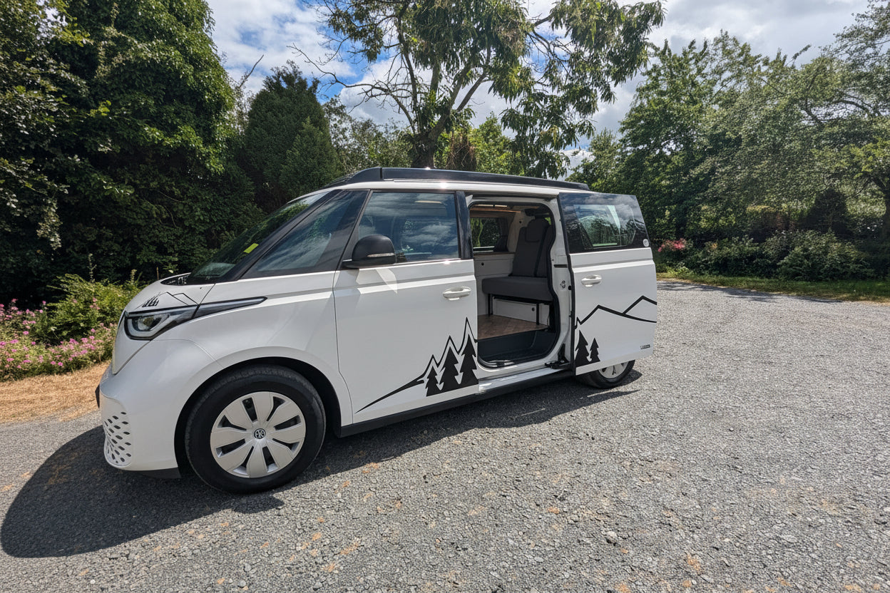 A white electric vw id buzz campervan with sliding side doors open, parked on a gravel surface outdoors with trees and a cloudy blue sky in the background. Black mountain and tree graphics decorate the side of the vehicle.