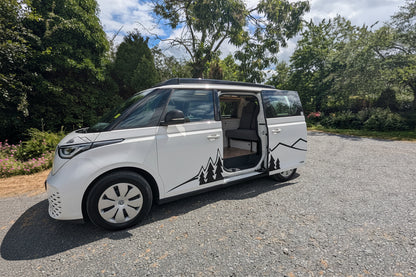 A white electric vw id buzz campervan with sliding side doors open, parked on a gravel surface outdoors with trees and a cloudy blue sky in the background. Black mountain and tree graphics decorate the side of the vehicle.