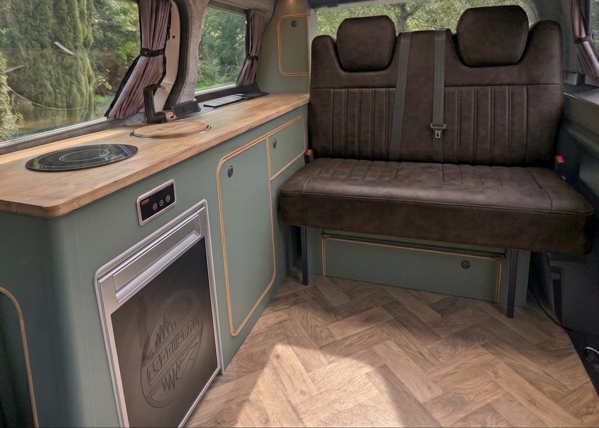 Rear seating area of a vw id buzz camper van interior with a brown upholstered bench, wood countertop, built-in stove and refrigerator, and a wood herringbone floor. Trees are visible through the windows and roof opening.