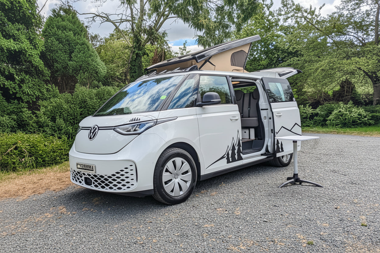 A white Volkswagen vw id buzz camper van parked on a gravel surface outdoors, with trees, bushes, and a blue sky with clouds in the background. The side door and pop-up roof are open.