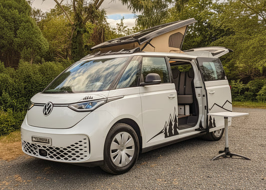 A white Volkswagen vw id buzz camper van parked on a gravel surface outdoors, with trees, bushes, and a blue sky with clouds in the background. The side door and pop-up roof are open.