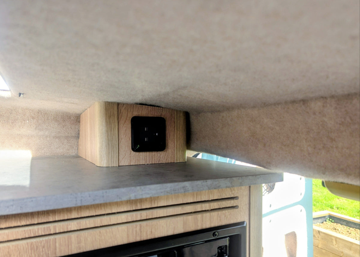 Close-up of a black multi-voltage power socket mounted on a custom wooden housing near the pop-top roof lining of the Kia PV5 Witley campervan.