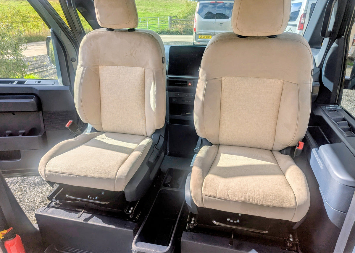 Front cabin view showing the adjustable upholstered driver and passenger swivel seats in matching beige fabric and suede inside the Kia PV5 Witley.