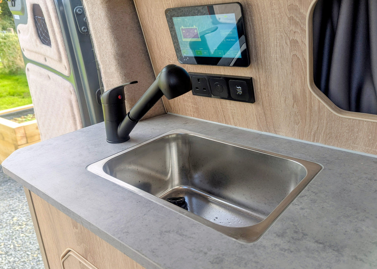 Close-up of the stainless steel kitchen sink, black gooseneck faucet, and digital system monitoring panel inside the Kia PV5 Witley electric campervan.