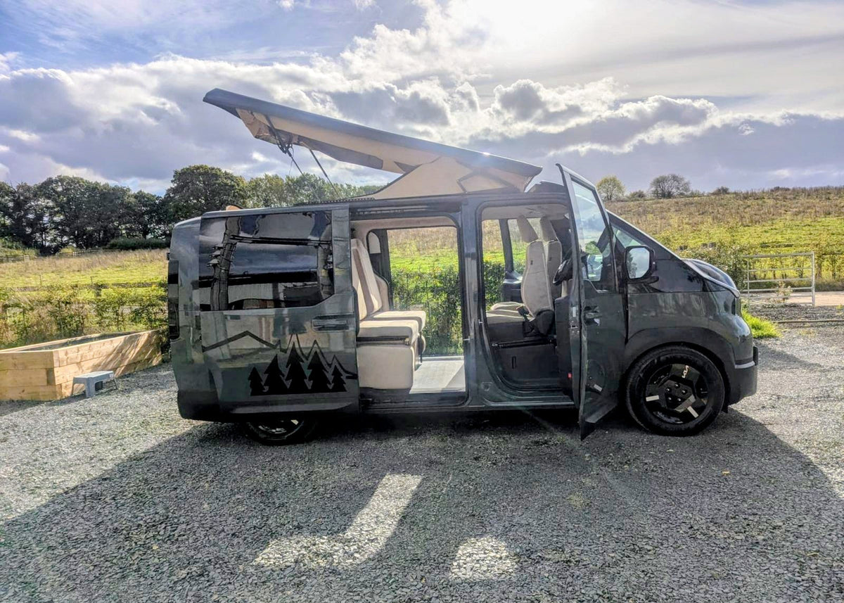 Side profile of the Kia PV5 Witley electric campervan with its pop-top roof elevated and side sliding door open in a rural setting, showcasing its compact camper layout.