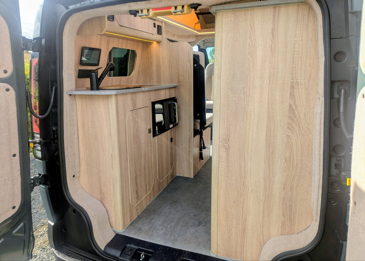 View from the open rear doors of the Kia PV5 Witley electric campervan highlighting the fitted kitchen counter, stainless steel sink, and custom wood cabinetry
