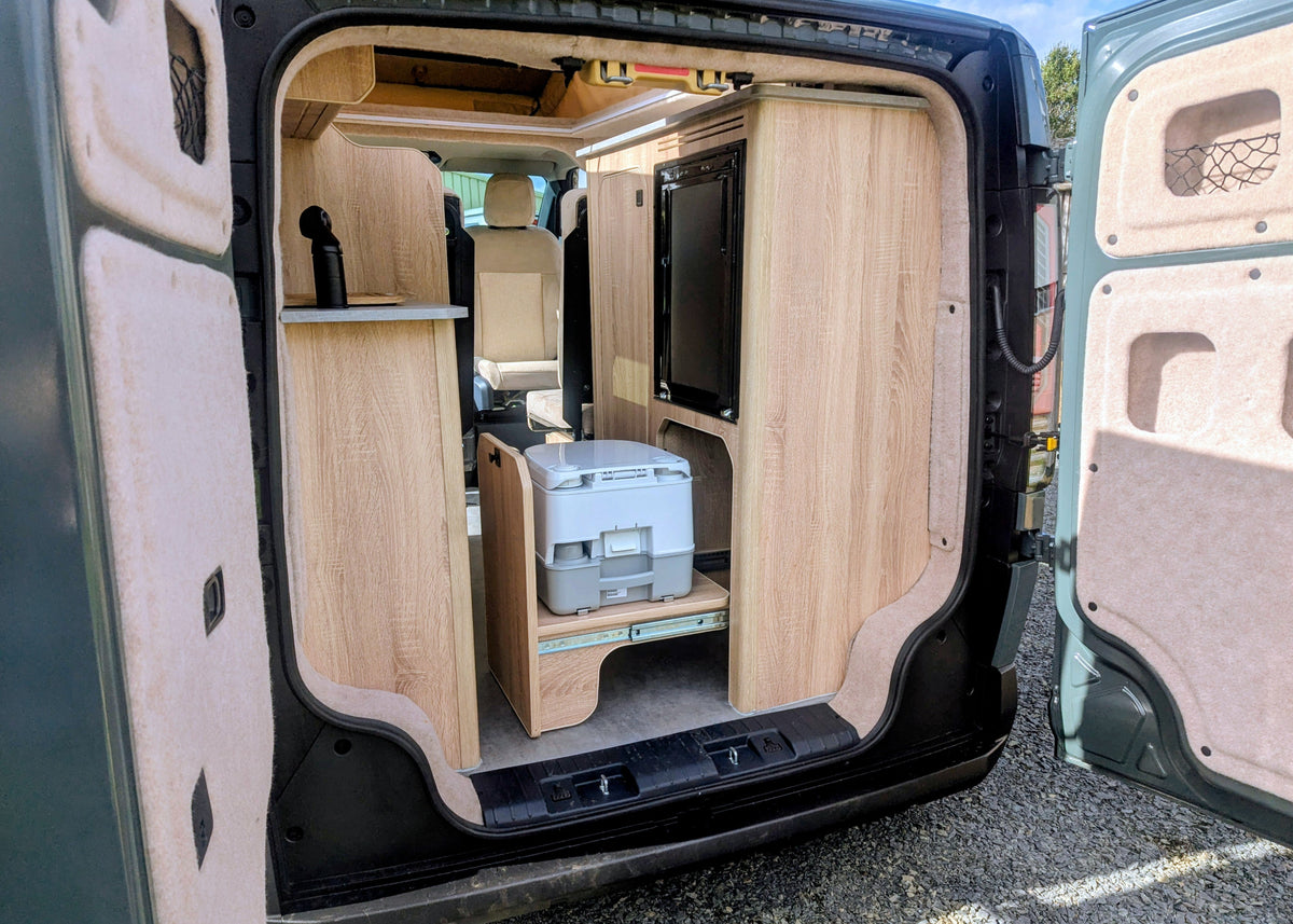 Rear storage garage area of the Kia PV5 Witley electric campervan featuring a custom pull-out slide holding a portable chemical toilet beneath the kitchen units.