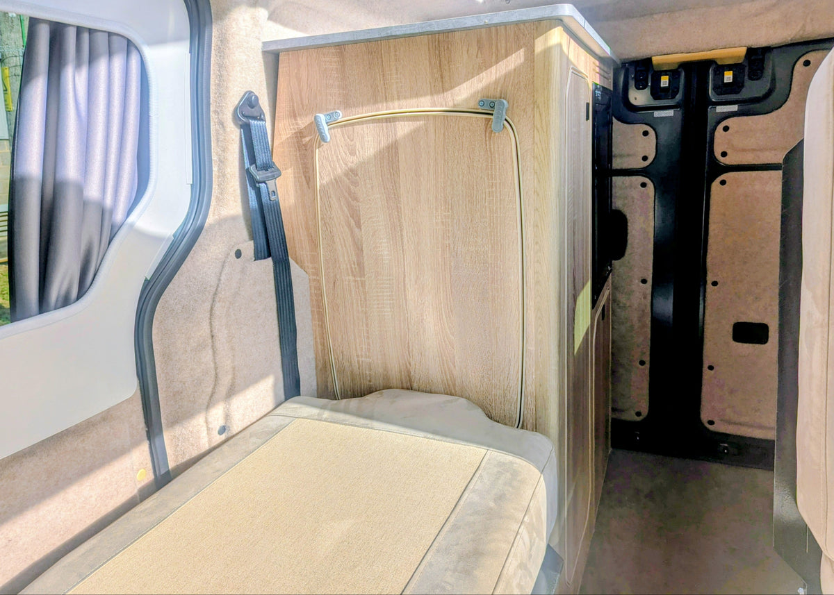 Interior view of the Kia PV5 Witley campervan showing the folded seat conversion into bed platform configuration next to the main kitchen cabinetry with intergrated table.