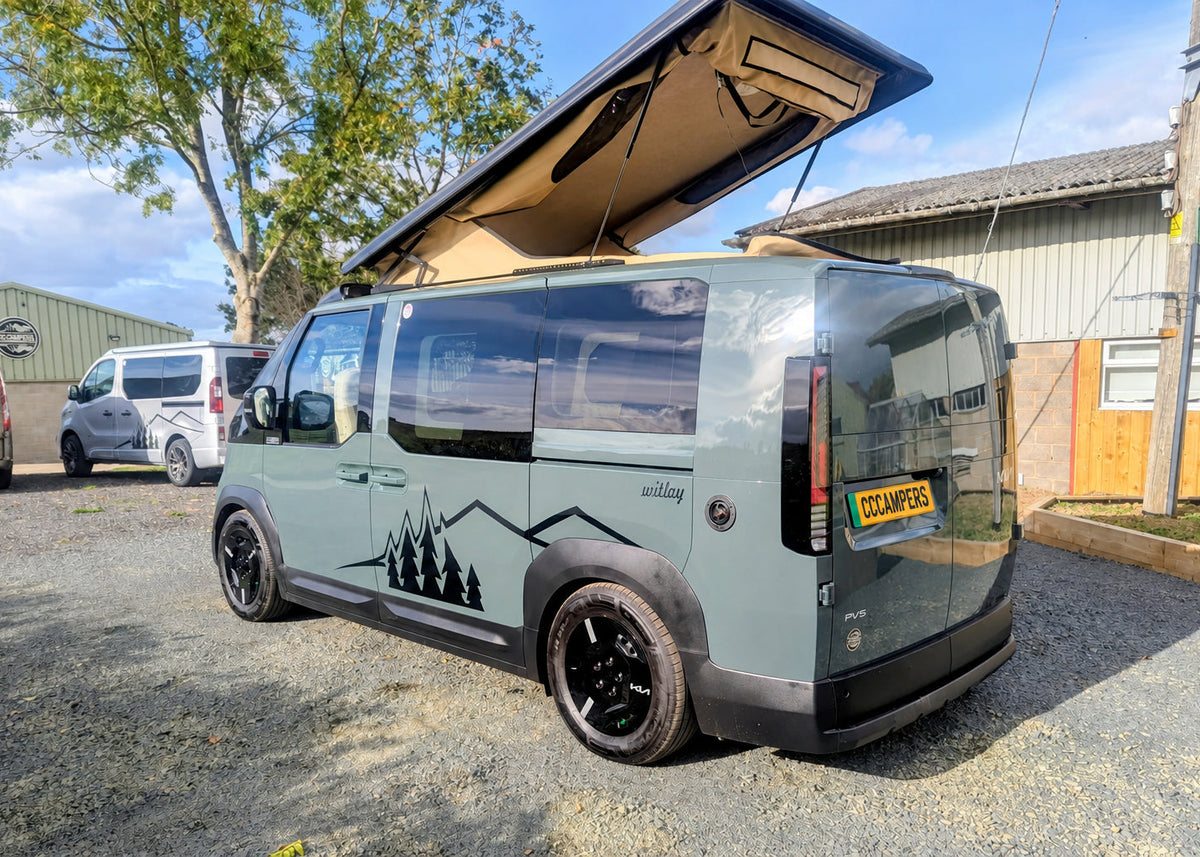 Rear three-quarter exterior view of the Kia PV5 Witley electric campervan showcasing the mountain side decal, custom plates, and raised roof.