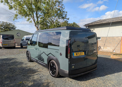 Rear three-quarter exterior perspective of the grey Kia PV5 Witley electric campervan showing the split rear doors and signature mountain graphics.