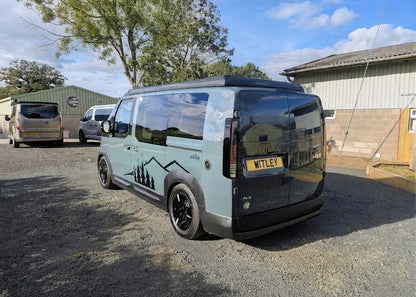 Rear three-quarter exterior view of the Kia PV5 Witley electric campervan conversion, featuring bespoke mountain graphic decals, black alloy wheels, and an elevated pop-top roof.