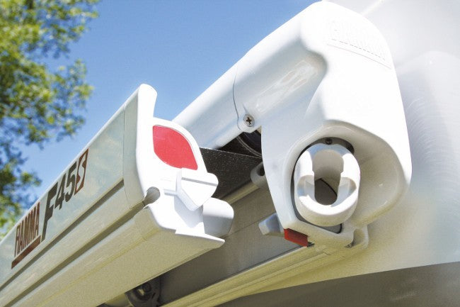Close-up of a white and gray retractable awning with a red release button, mounted on a vehicle or structure outdoors under a blue sky.