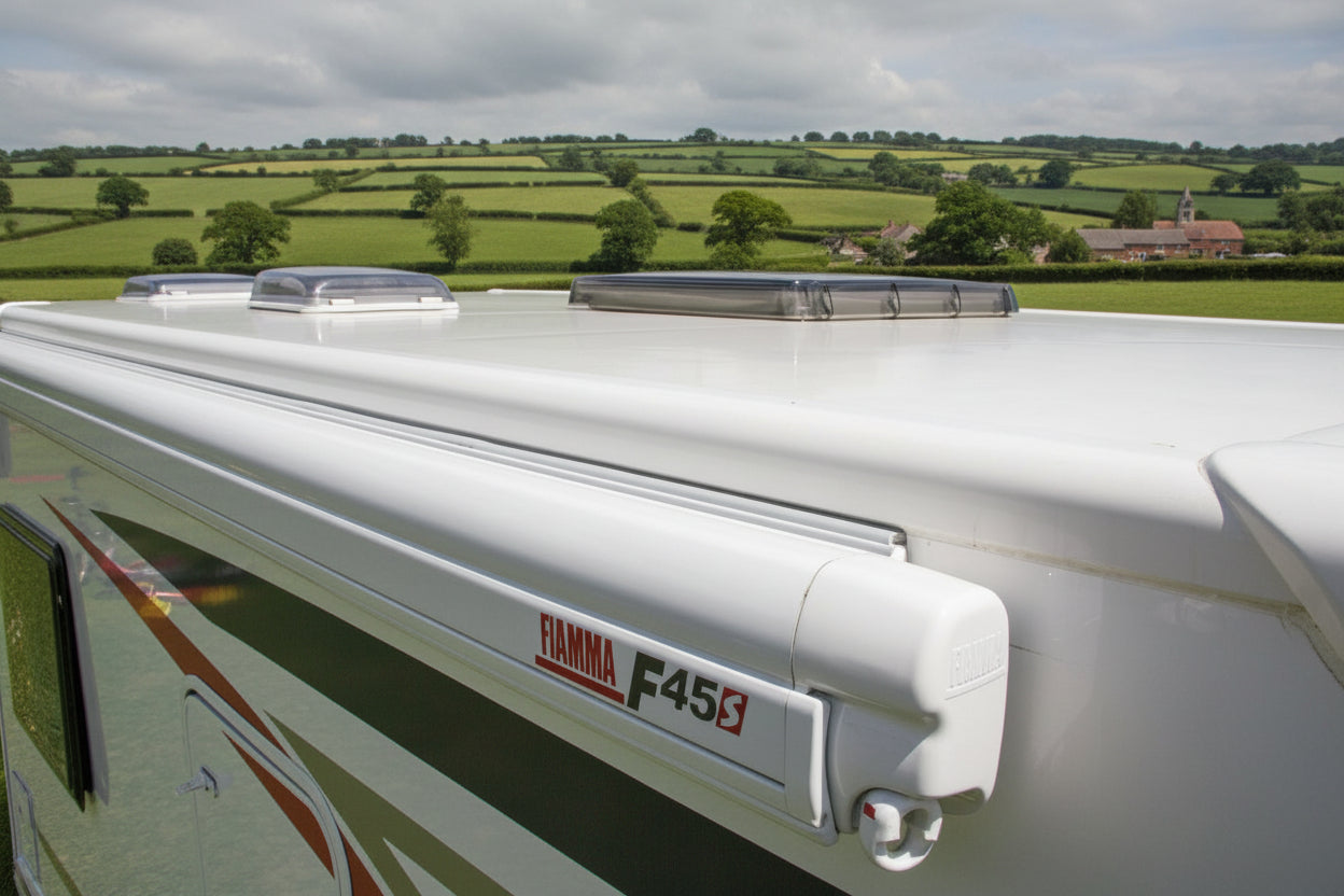 Close-up of a white caravan or motorhome with FIAMMA F45s side awning, set against a background of green rural fields under a cloudy sky.
