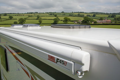 Close-up of a white caravan or motorhome with FIAMMA F45s side awning, set against a background of green rural fields under a cloudy sky.