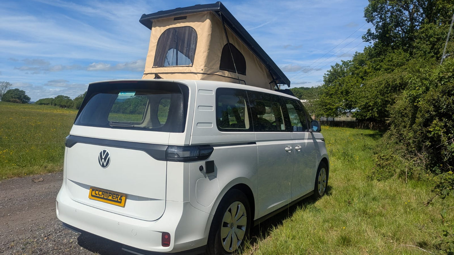 White Volkswagen van with a rooftop tent parked on a gravel road next to a grassy area with trees.