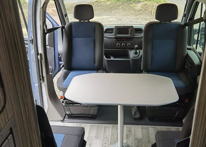 A Motorhome interior with two blue and gray seats, a fold-out white table in the center, and an open side door showing the vehicle's dashboard and outdoor view.