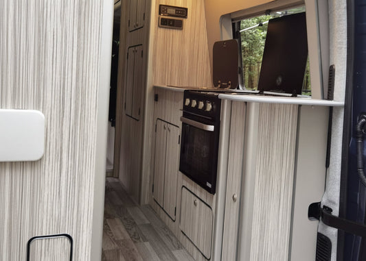 The rear kitchen area of a motorhome or camper van, featuring light wood grain cabinets, a black built-in oven, a small countertop stove, and a window looking out onto trees.